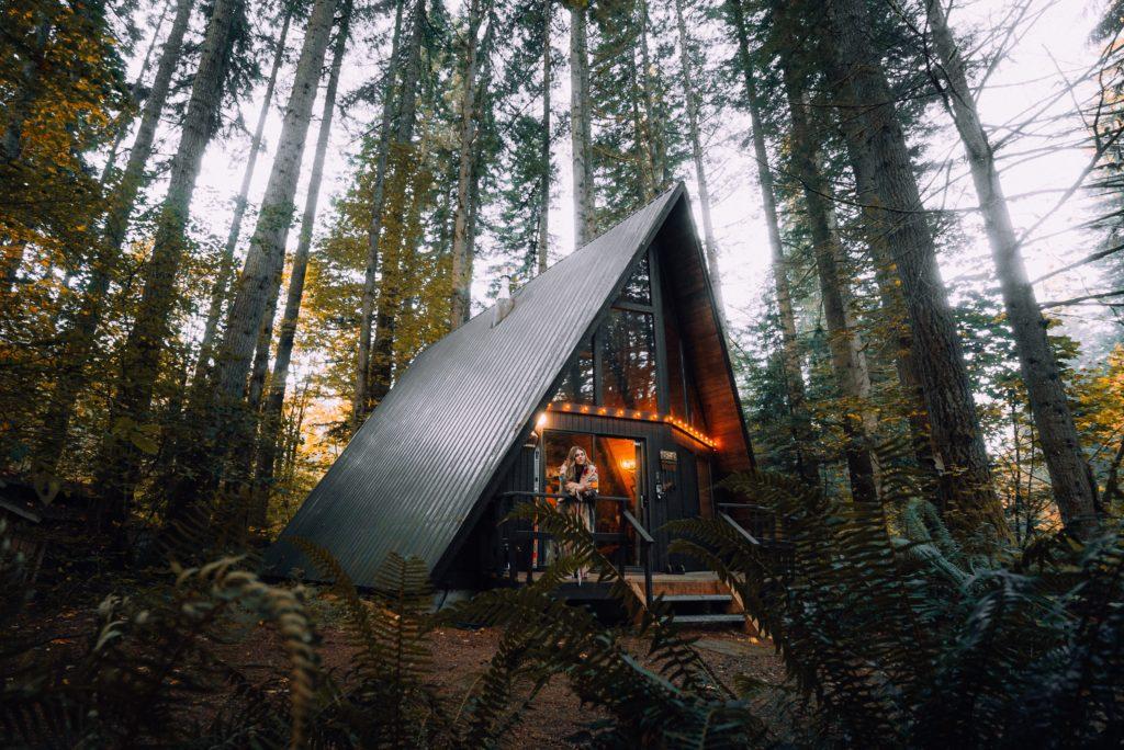 A modern A-frame cabin nestled among tall trees in a lush forest, featuring warm lighting and a welcoming entrance, ideal for a cozy getaway in nature.