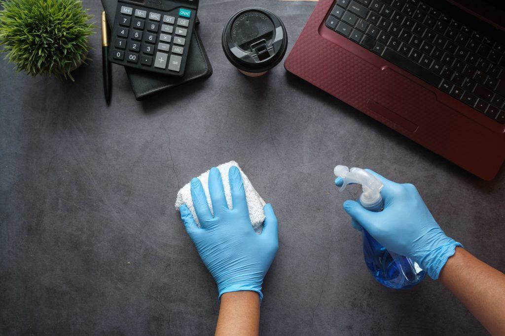 A person wearing blue gloves cleaning a desk with a spray bottle and cloth, surrounded by a calculator, coffee cup, and laptop, emphasizing the importance of maintaining a clean workspace.
