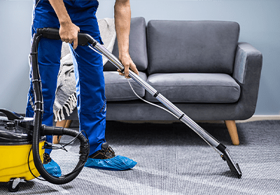 A professional cleaner in blue scrubs using a vacuum cleaner on a carpet near a gray sofa, wearing protective shoe covers for cleanliness.