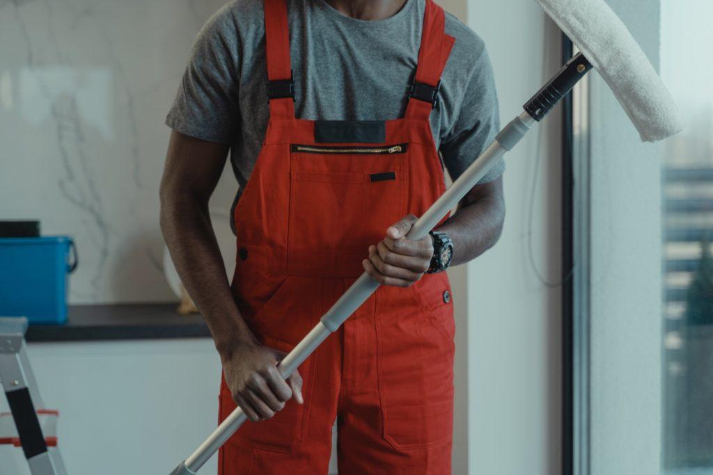 A person wearing red overalls and a gray shirt holds a long cleaning tool, preparing to clean a window or surface.