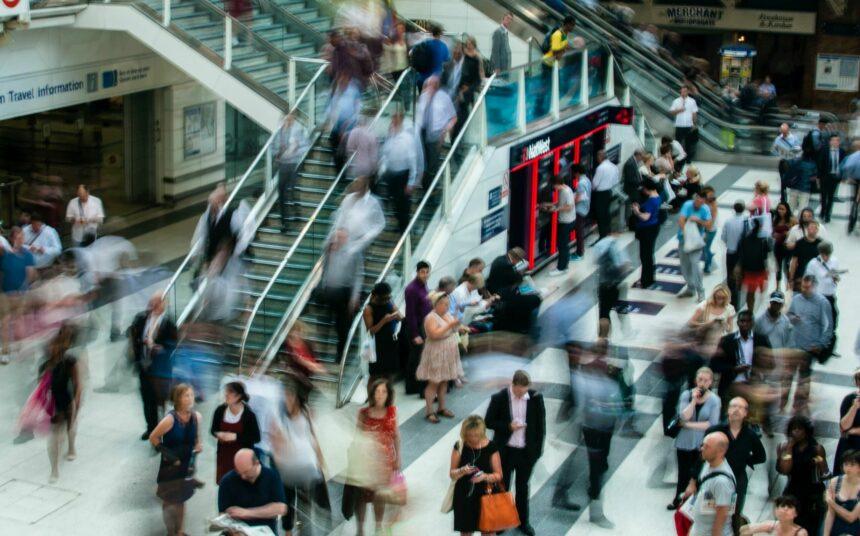 Busy public space with people commuting and interacting, featuring an escalator and a vibrant crowd in motion.