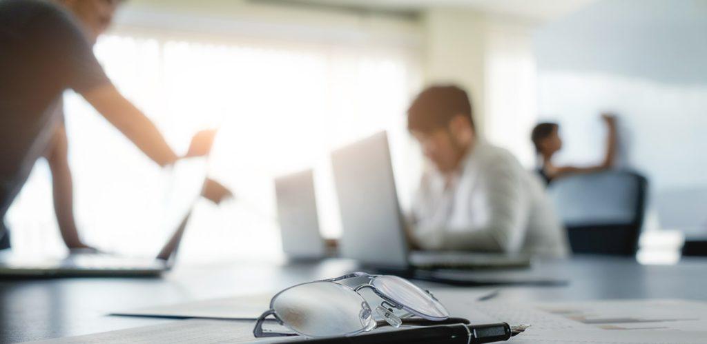 A blurred office setting with two individuals working on laptops, while a pair of glasses rests on a desk in the foreground, highlighting a focused work environment.