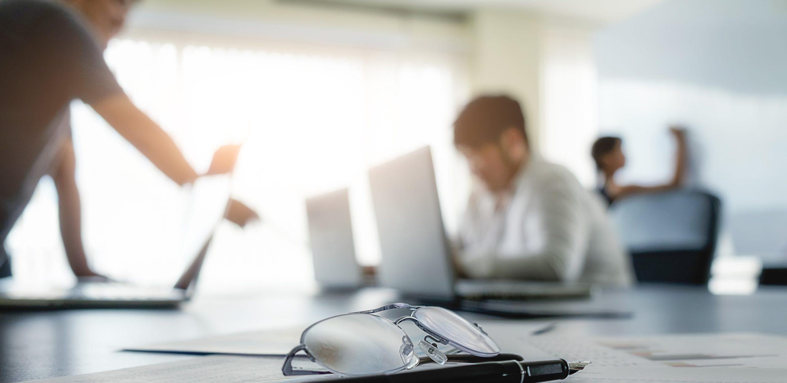 A blurred office setting with two individuals working on laptops, while a pair of glasses rests on a desk in the foreground, highlighting a focused work environment.