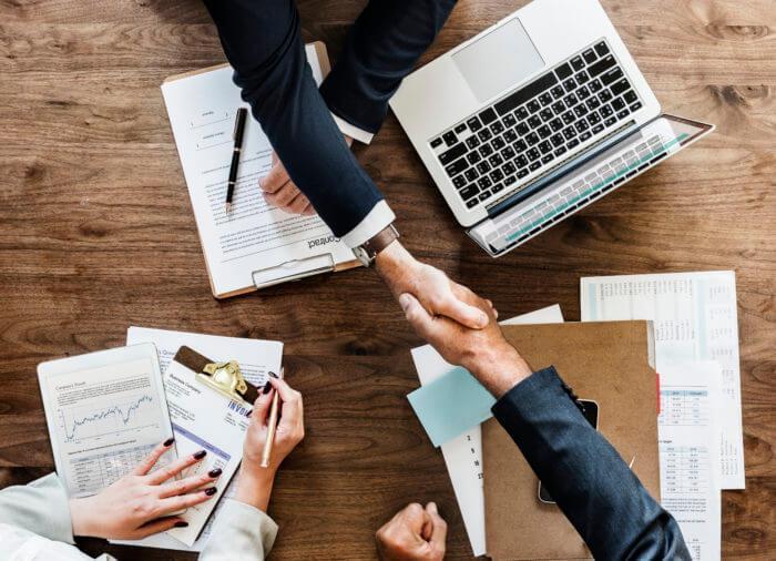 Business professionals shaking hands over a desk with a laptop and paperwork, symbolizing a successful partnership or agreement.
