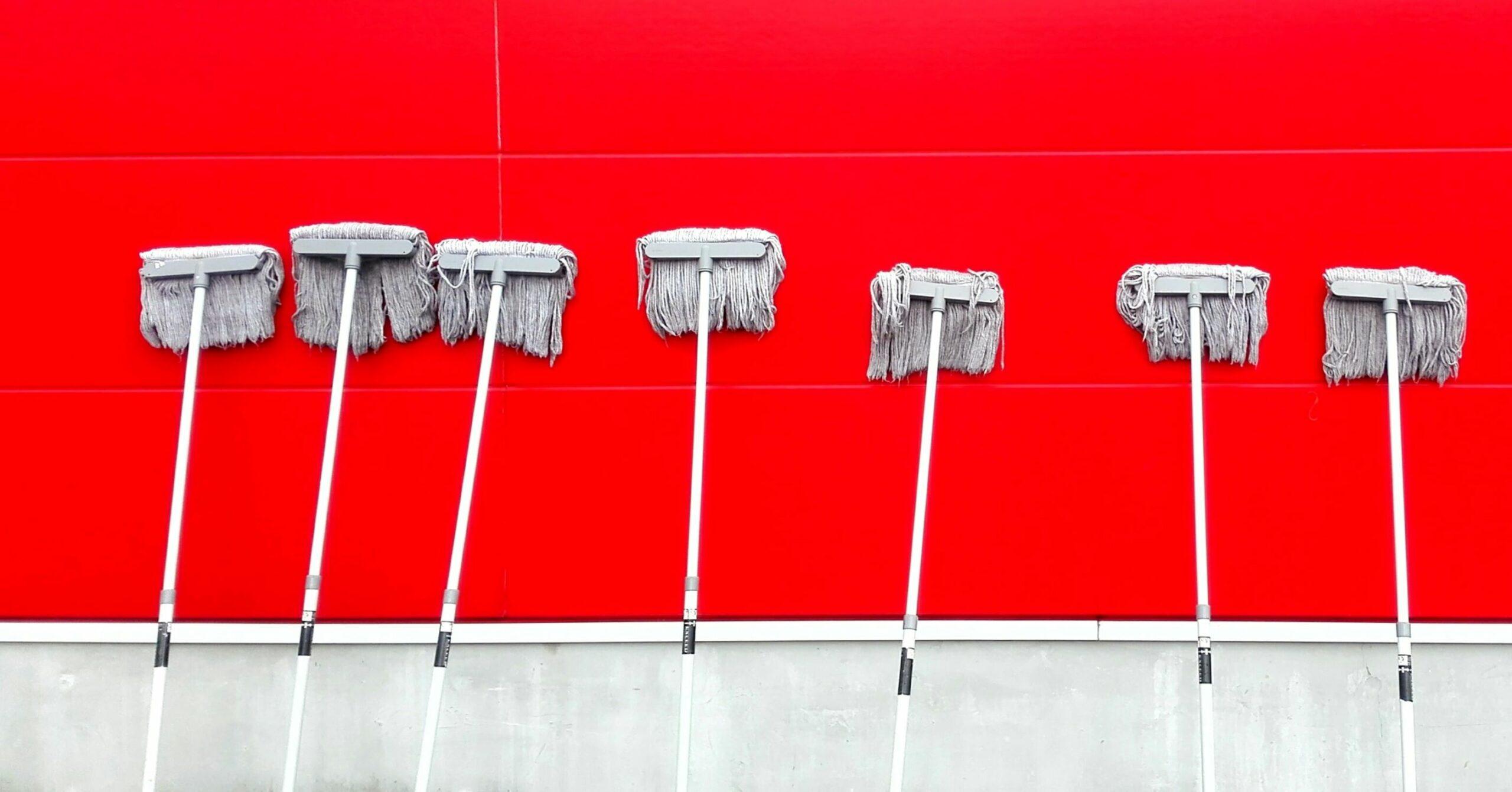 Five cleaning mops lined up against a bright red wall, showcasing different sizes and styles for effective floor maintenance.