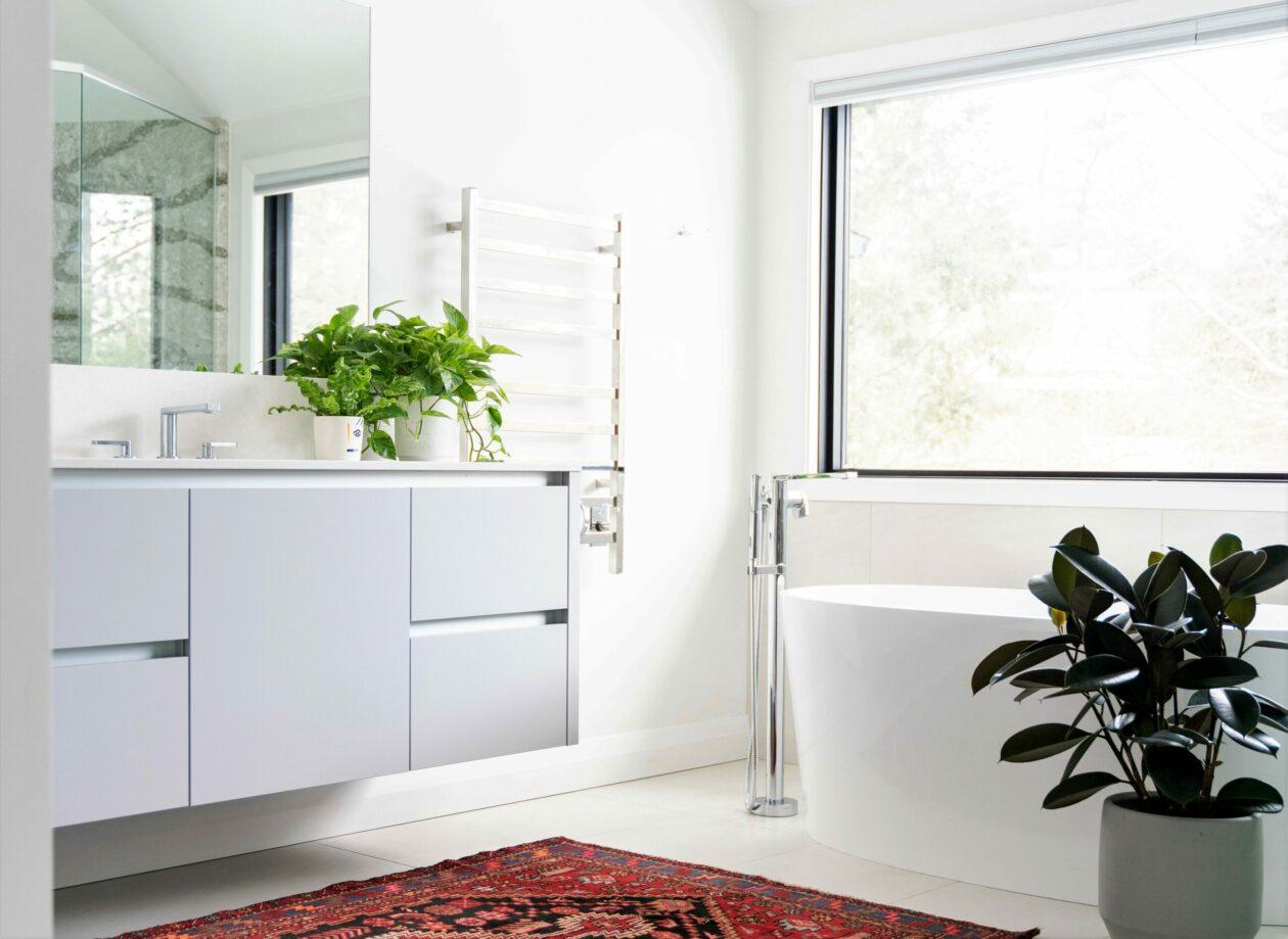 Modern bathroom interior featuring a sleek white vanity, large window for natural light, a freestanding bathtub, and decorative plants, creating a stylish and serene atmosphere.