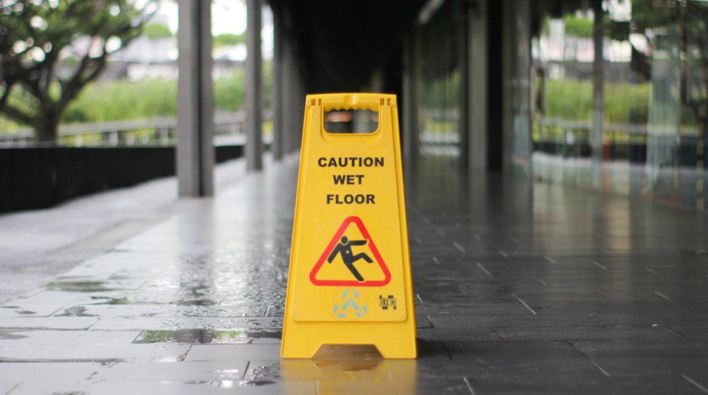 Yellow caution sign warning of wet floor in a hallway, indicating potential slip hazards for pedestrians.