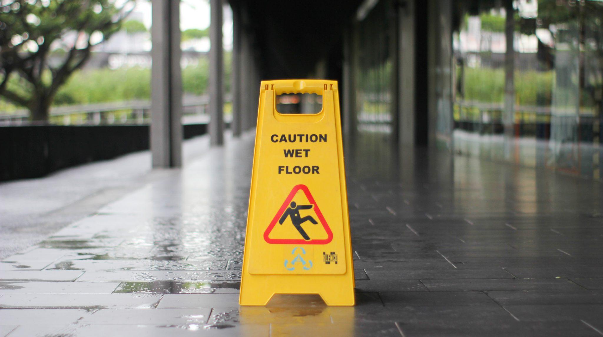 Yellow caution sign warning of wet floor in a hallway, indicating potential slip hazards for pedestrians.