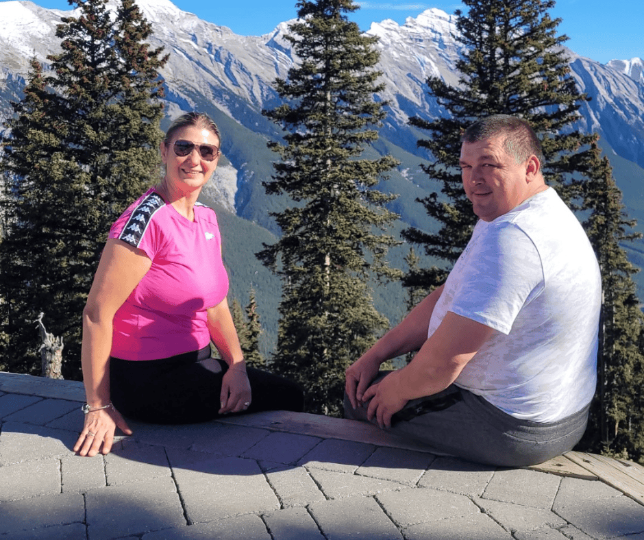 A man and a woman sitting on a wooden platform with a scenic mountain backdrop, surrounded by tall evergreen trees. The woman, wearing a pink athletic shirt and sunglasses, smiles at the camera, while the man, in a white t-shirt, looks back towards the camera, both enjoying a sunny day outdoors.