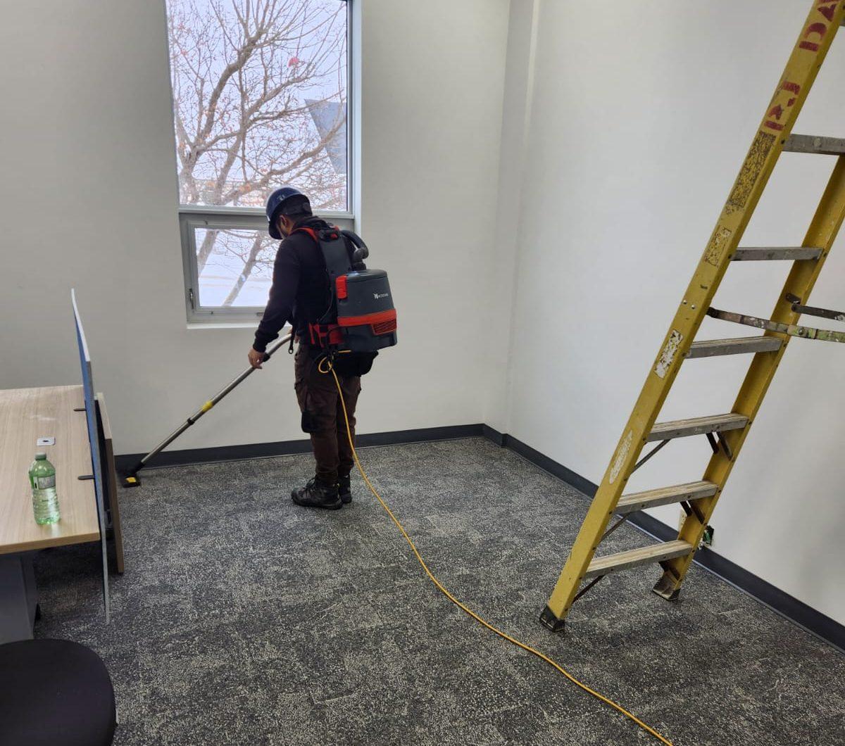 A professional cleaner using a backpack vacuum in a modern office space, with a ladder and a window in the background. The image showcases the cleaning process on carpeted flooring, emphasizing cleanliness and maintenance in a workspace environment.